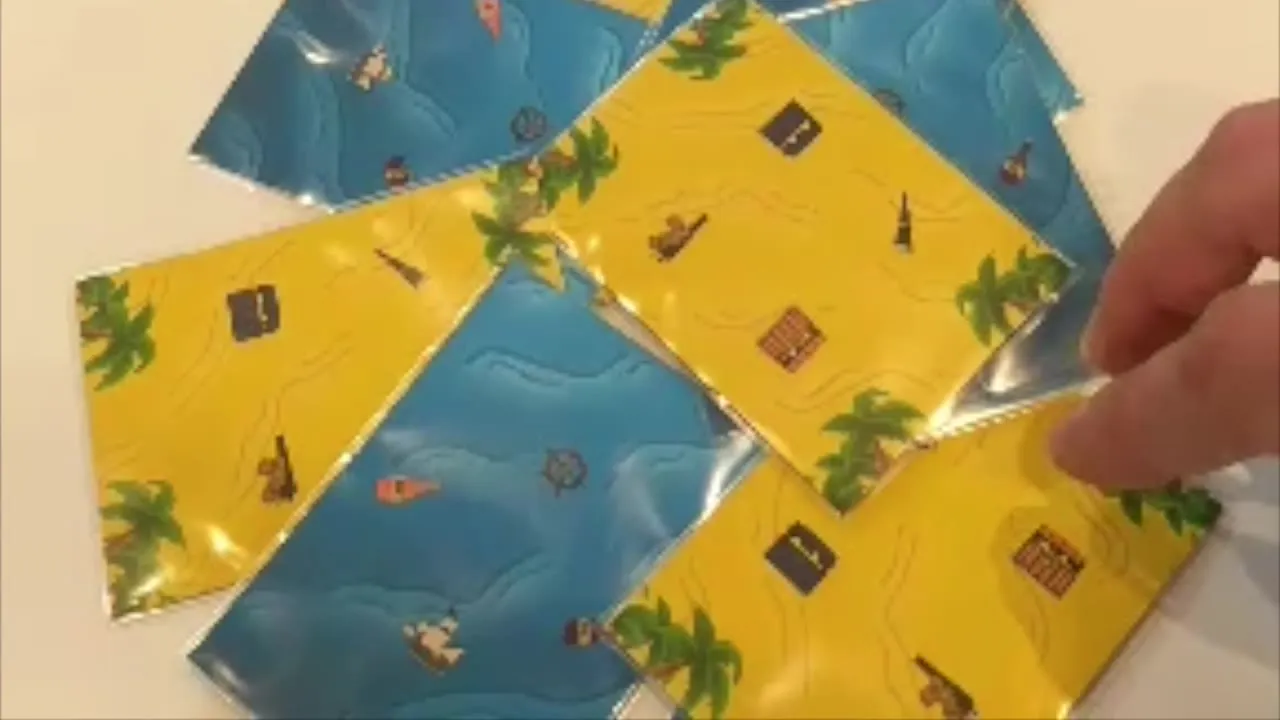 A close overhead view of six sleeved cards overlapping on a table, sand and ocean faces at angles, a hand entering from the right